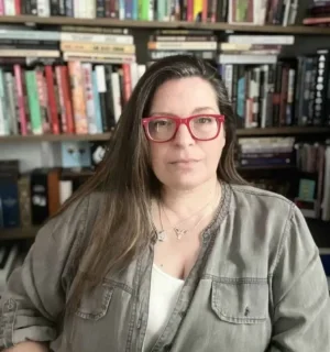 Image of Karla Strand. Strand is wearing red glasses and posing in front of several bookshelves filled with colorful books and arranged at various angles.